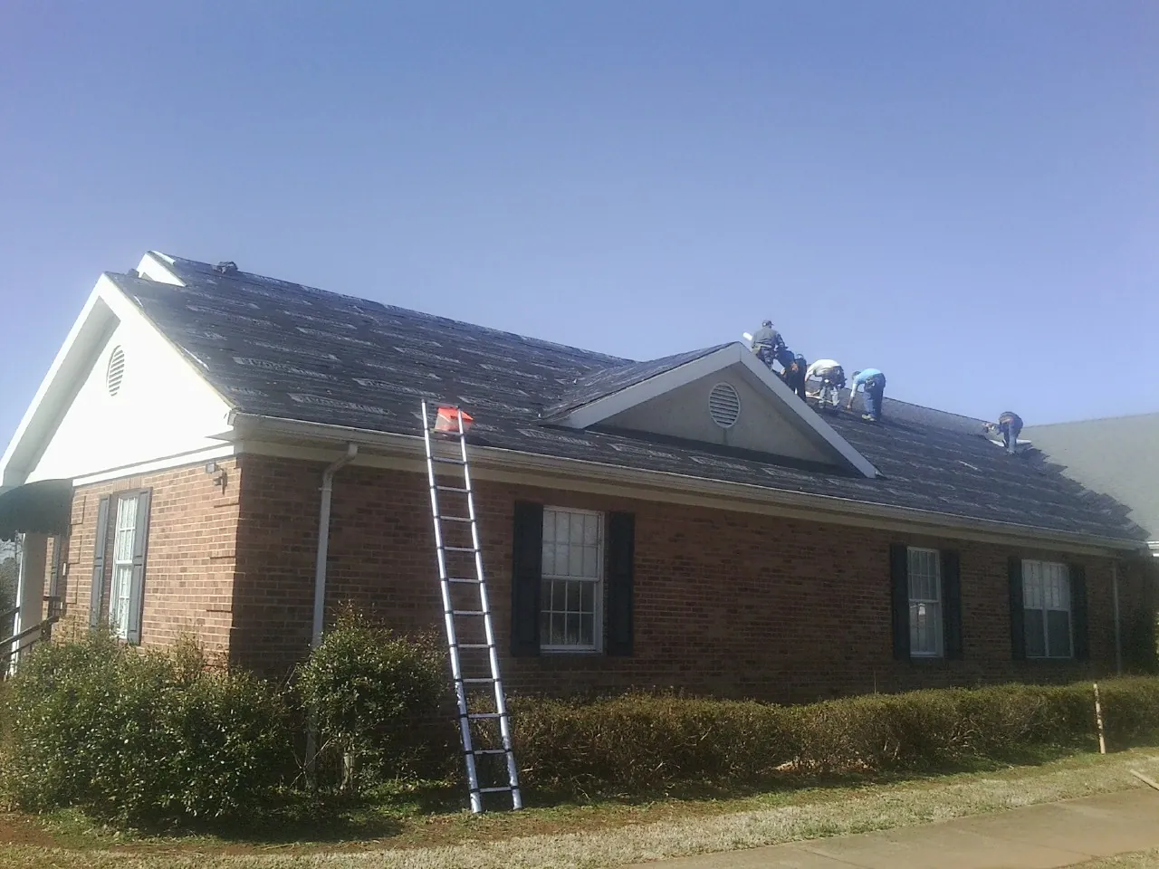 Crew tearing shingles off a brick ranch roof with a ladder set — Metal Roof Repair work by our crew in Weogufka