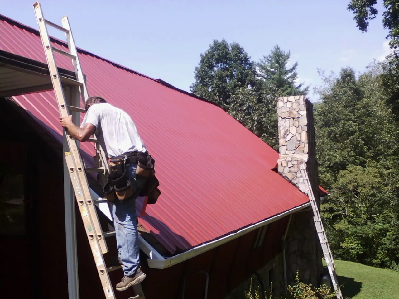 Roofer on a ladder beside a red metal roof and stone chimney — Soffit & Fascia Repair by Roofing Brothers in Weogufka